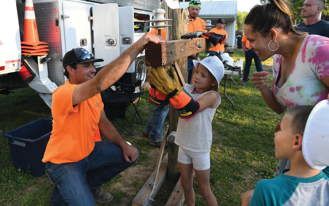 Journeyman Lineman Tyler Rhoades cheers on a young lineworker-in-training as she works to thread a nut on the bolt of a crossarm while wearing lineman’s rubber gloves and gear.